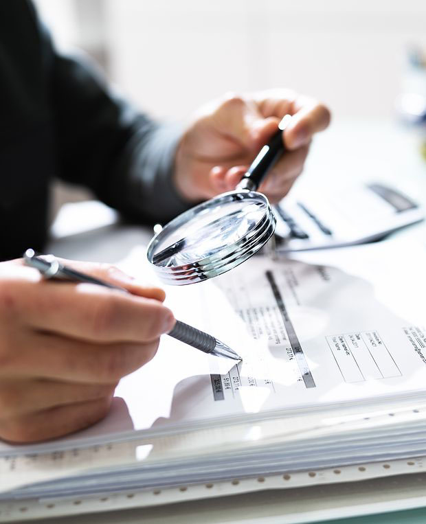 Person holding magnifying glass over audit paperwork.